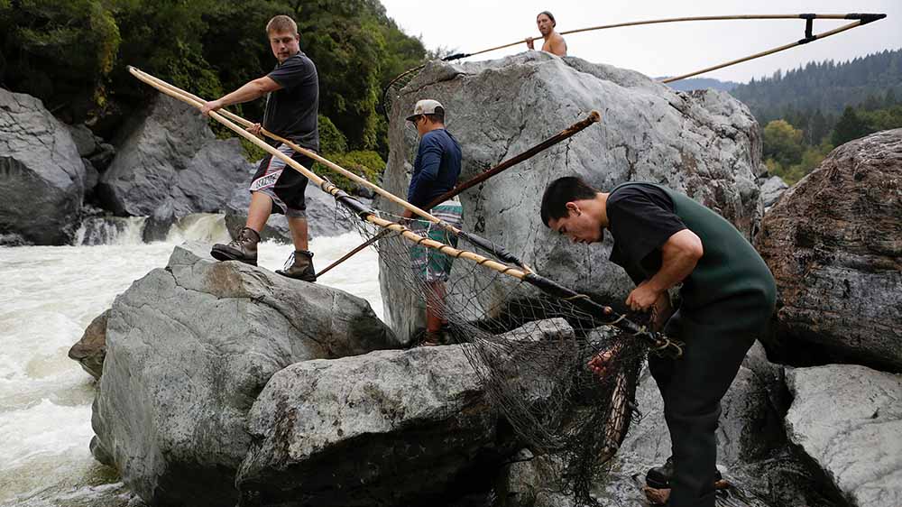Four men stand on a rocky beach working on traditional fishing equipment and nets.