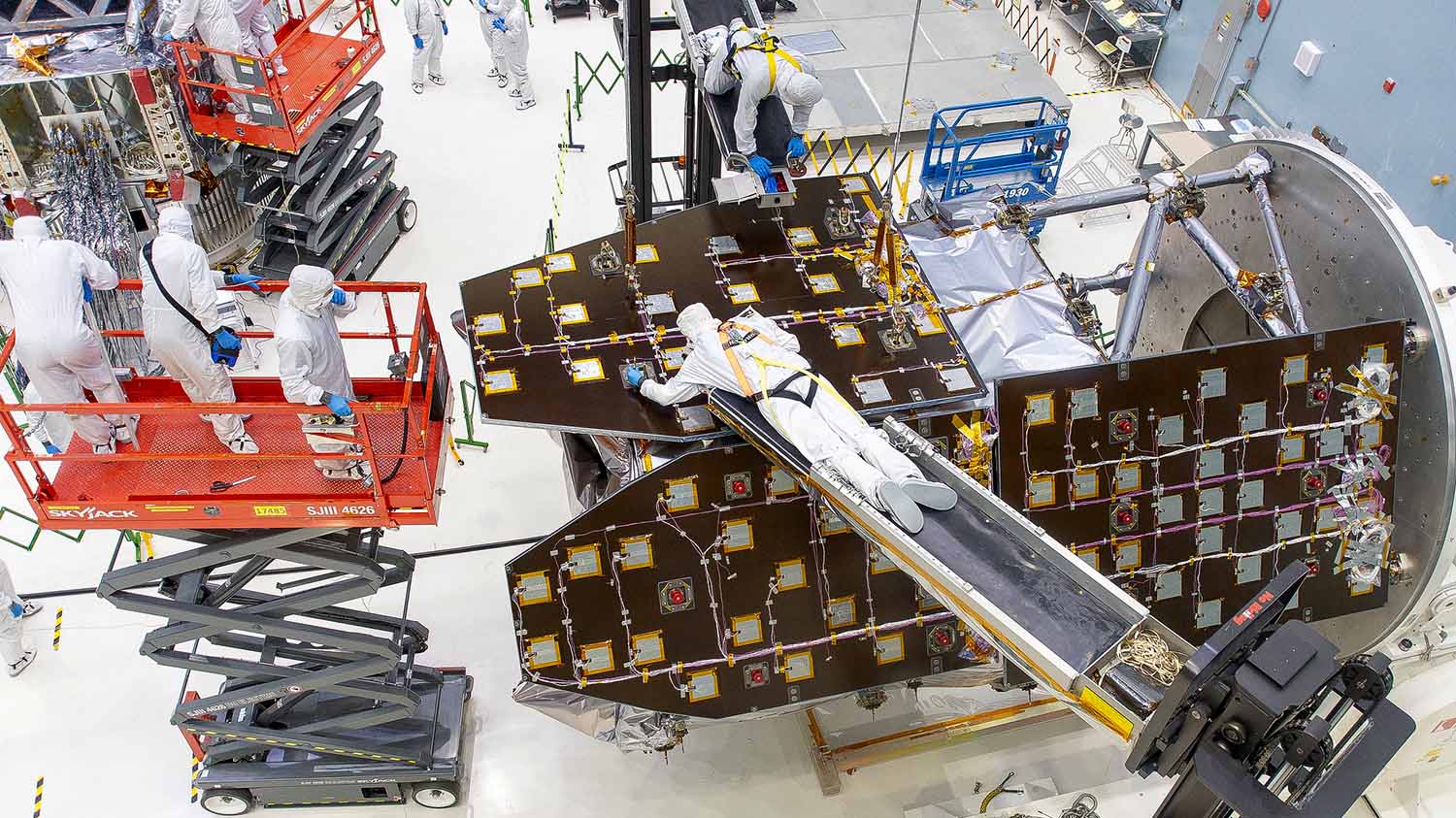 People in coveralls and gloves work on a space telescope that is under construction.