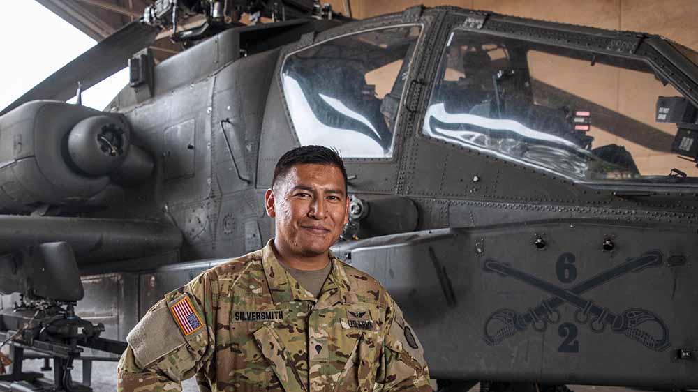 A man in a US army uniform poses in front of a military helicopter.