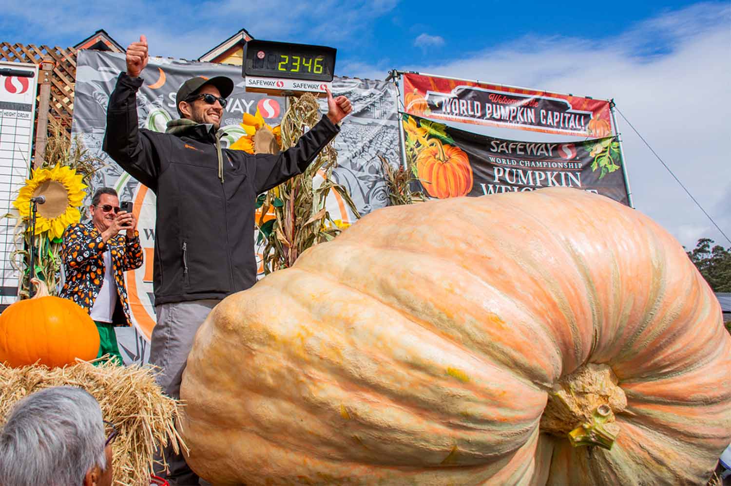 A giant pumpkin is on a scale that reads 2,346 pounds. Behind it, Brandon Dawson holds up his arms and gives two thumbs up.