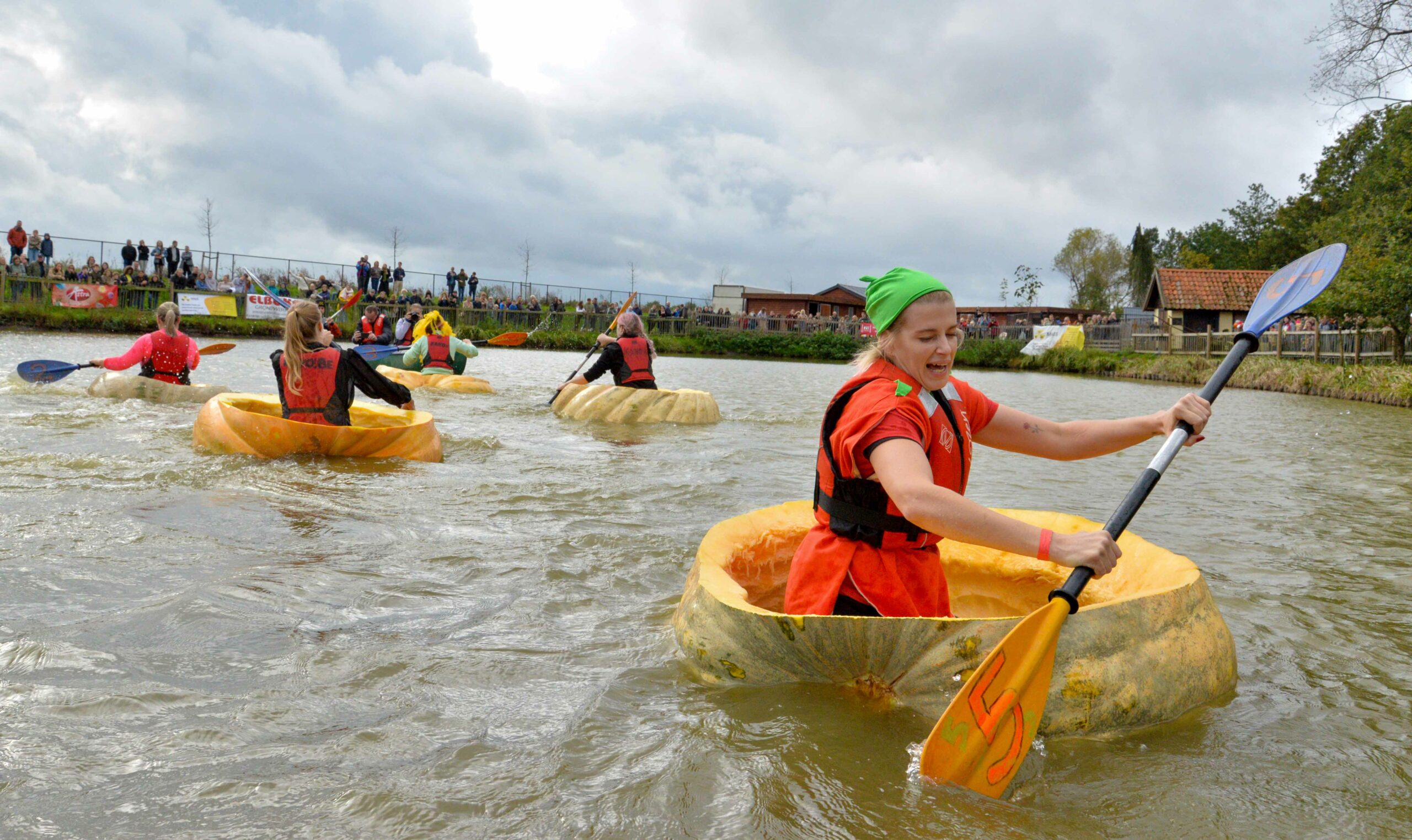 Five women each sit inside a hollowed out giant pumpkin and paddle on a body of water.