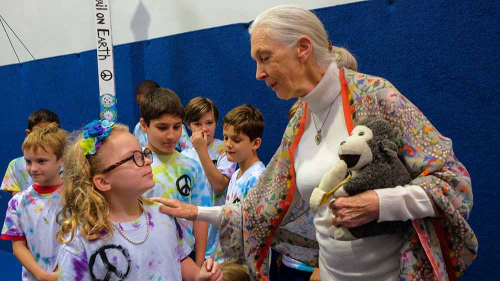 Older Jane Goodall holds a stuffed chimpanzee and talks to a young girl in a classroom or gym while other children are in the background.