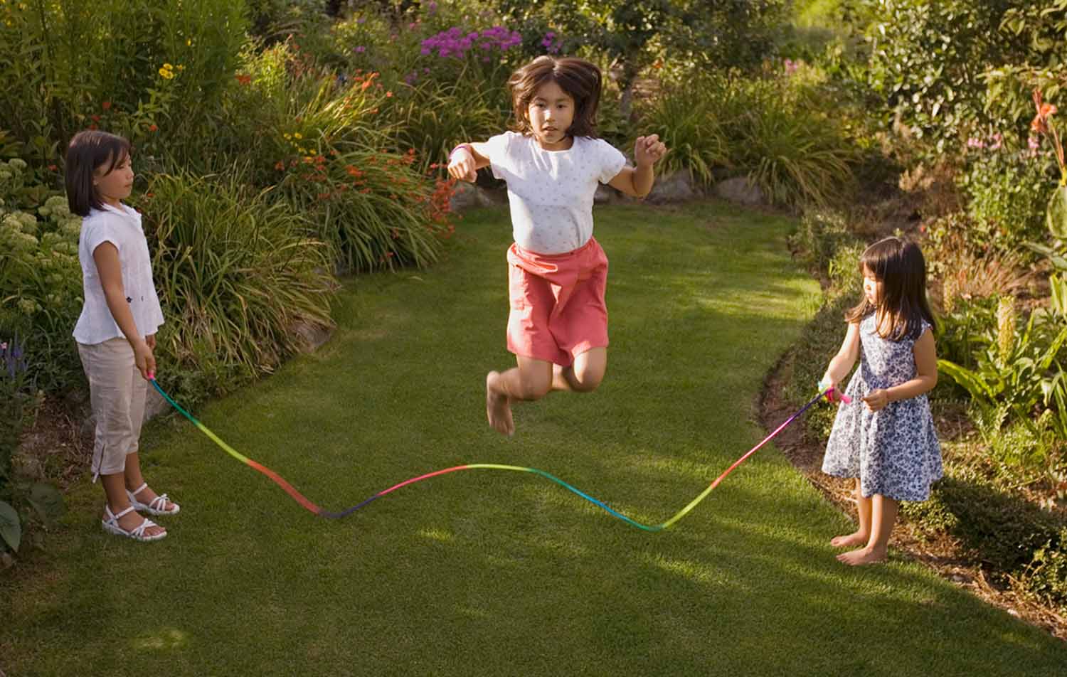 Three girls play jump rope on a lawn with a landscaped garden in the background.