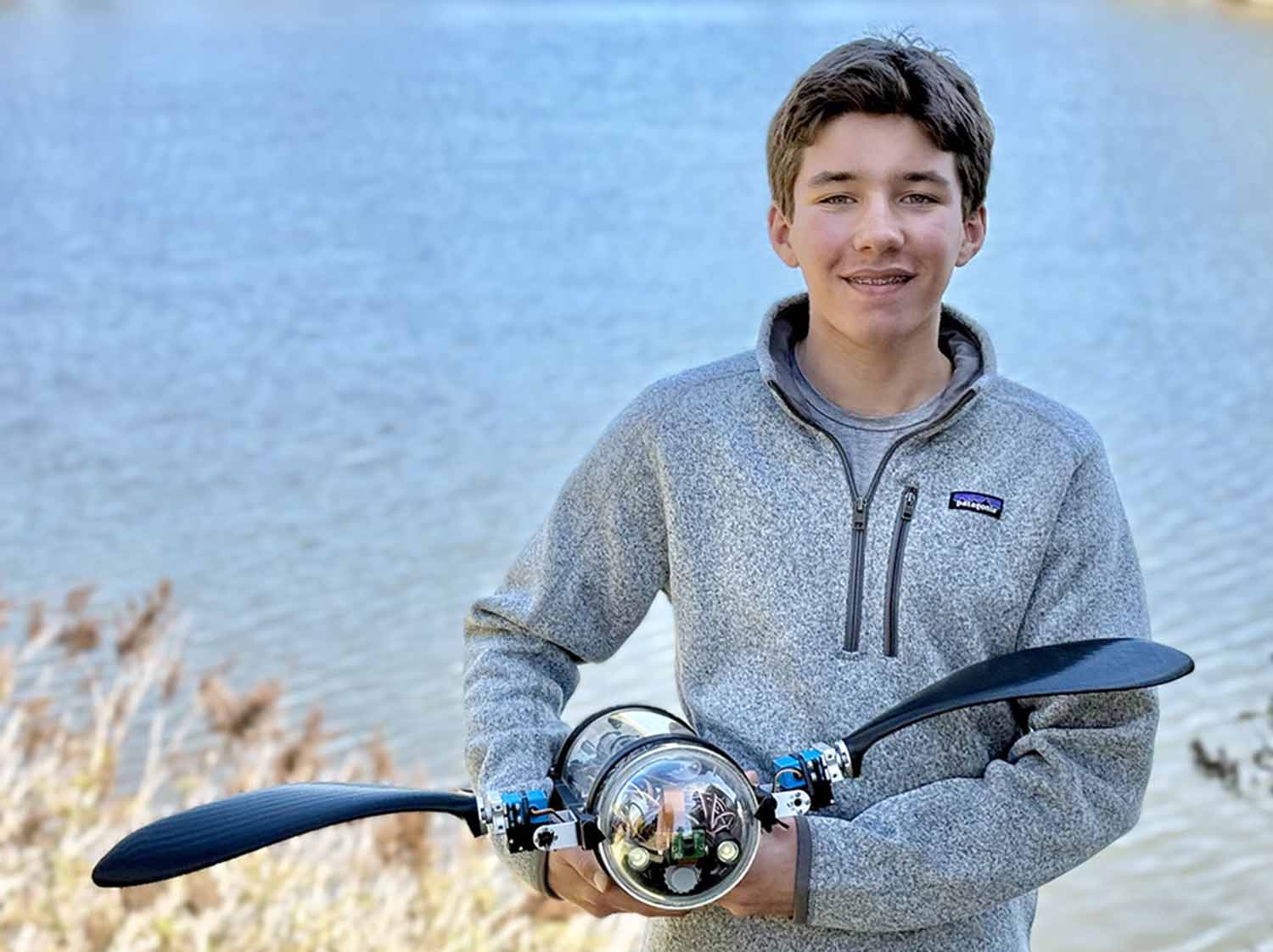 Evan Budz stands in front of a body of water and poses while holding a robot with flippers that resemble that of a sea turtle.