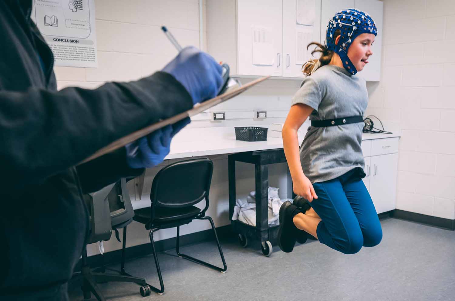 A child with equipment attached to her head and torso jumps in the air as an adult takes notes on a clipboard.