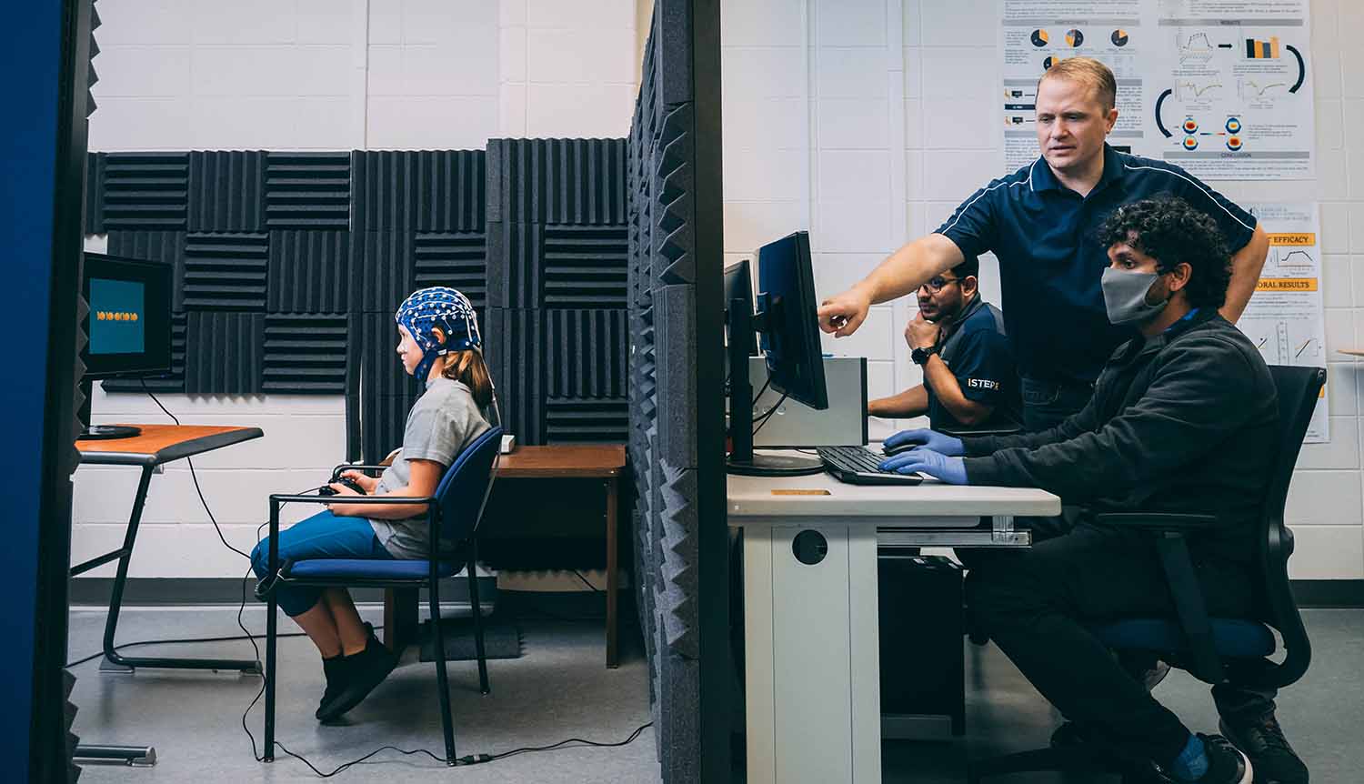 A child with electronic equipment attached to her head does a computer task while adults on the other side of a wall look at a computer screen.