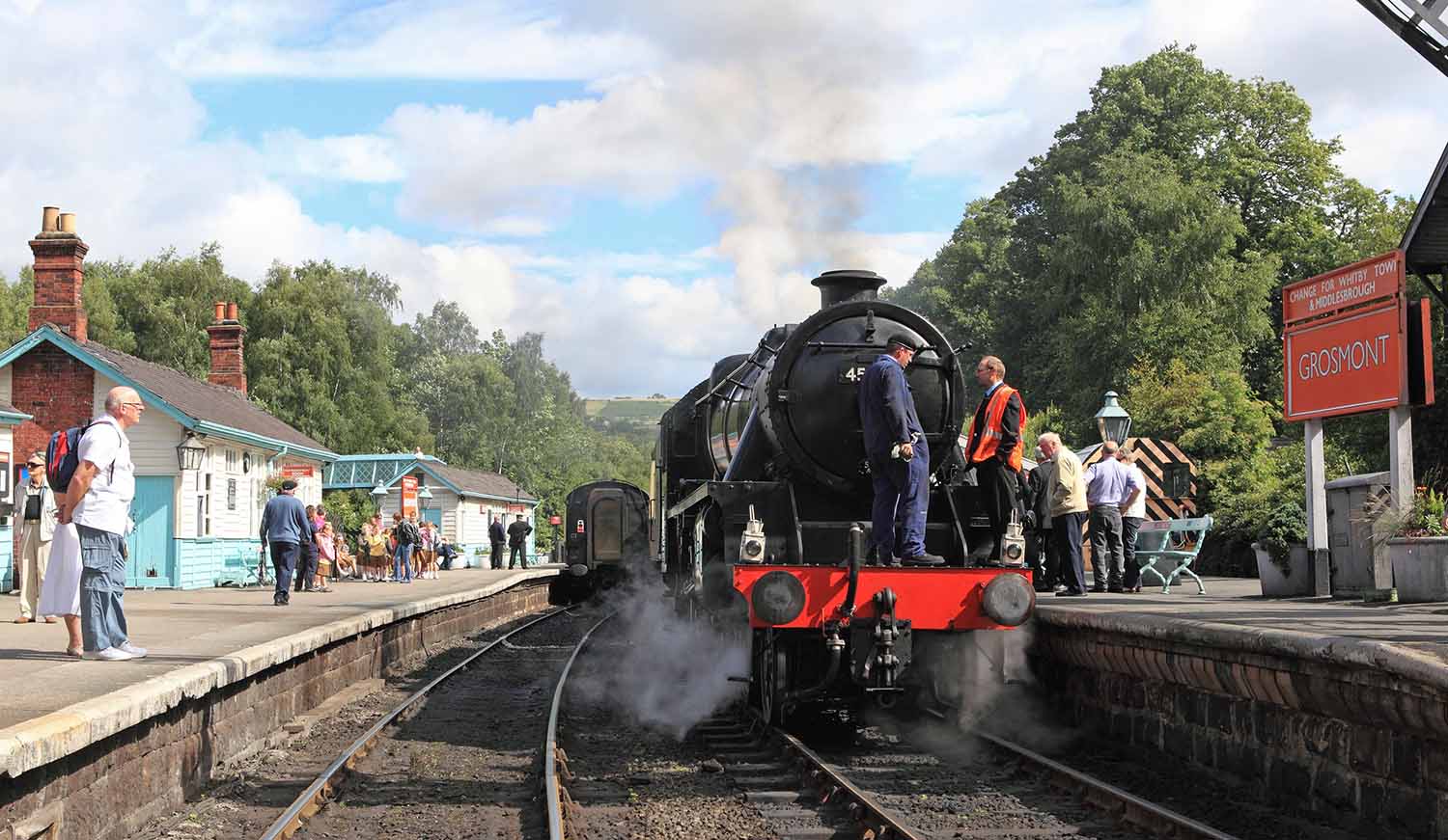 Two men stand on the front of an idle train at the Grosmont Station while others stand on the platform.
