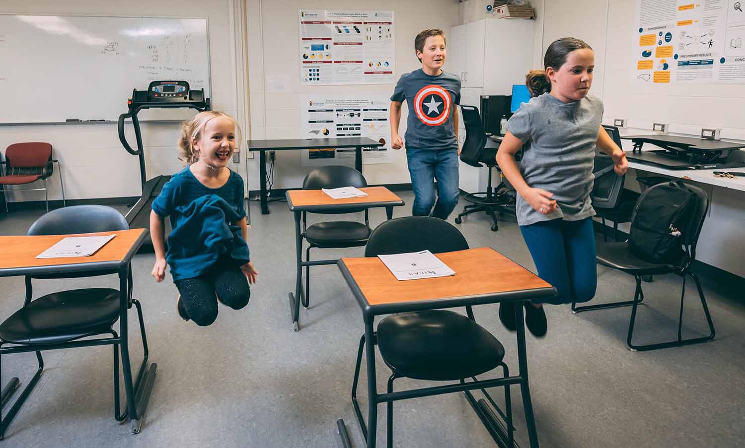 Three children jump in the air next to desks in a room that is set up like a classroom.