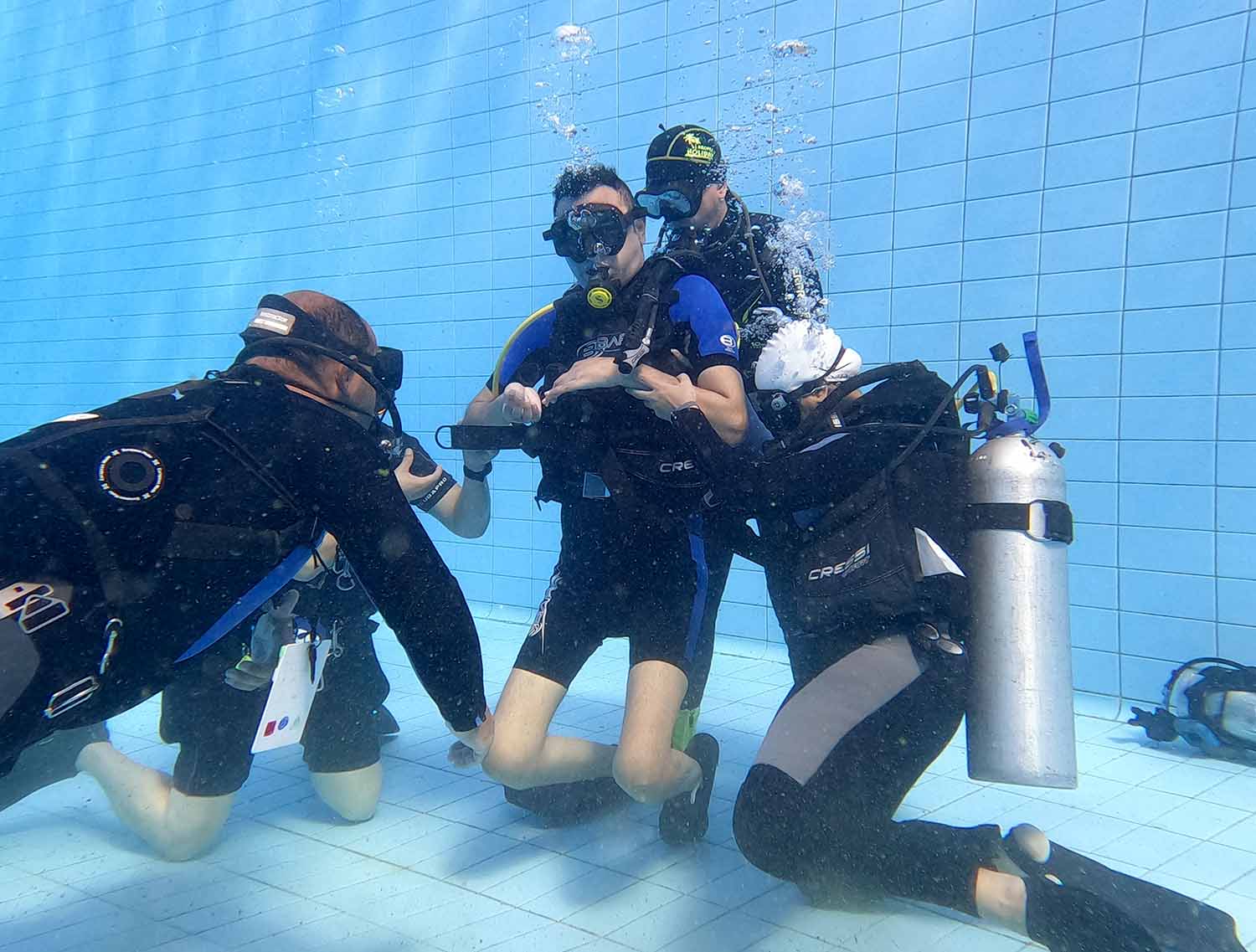 A man in scuba gear is at the bottom of a pool being assisted by three other men in scuba gear.