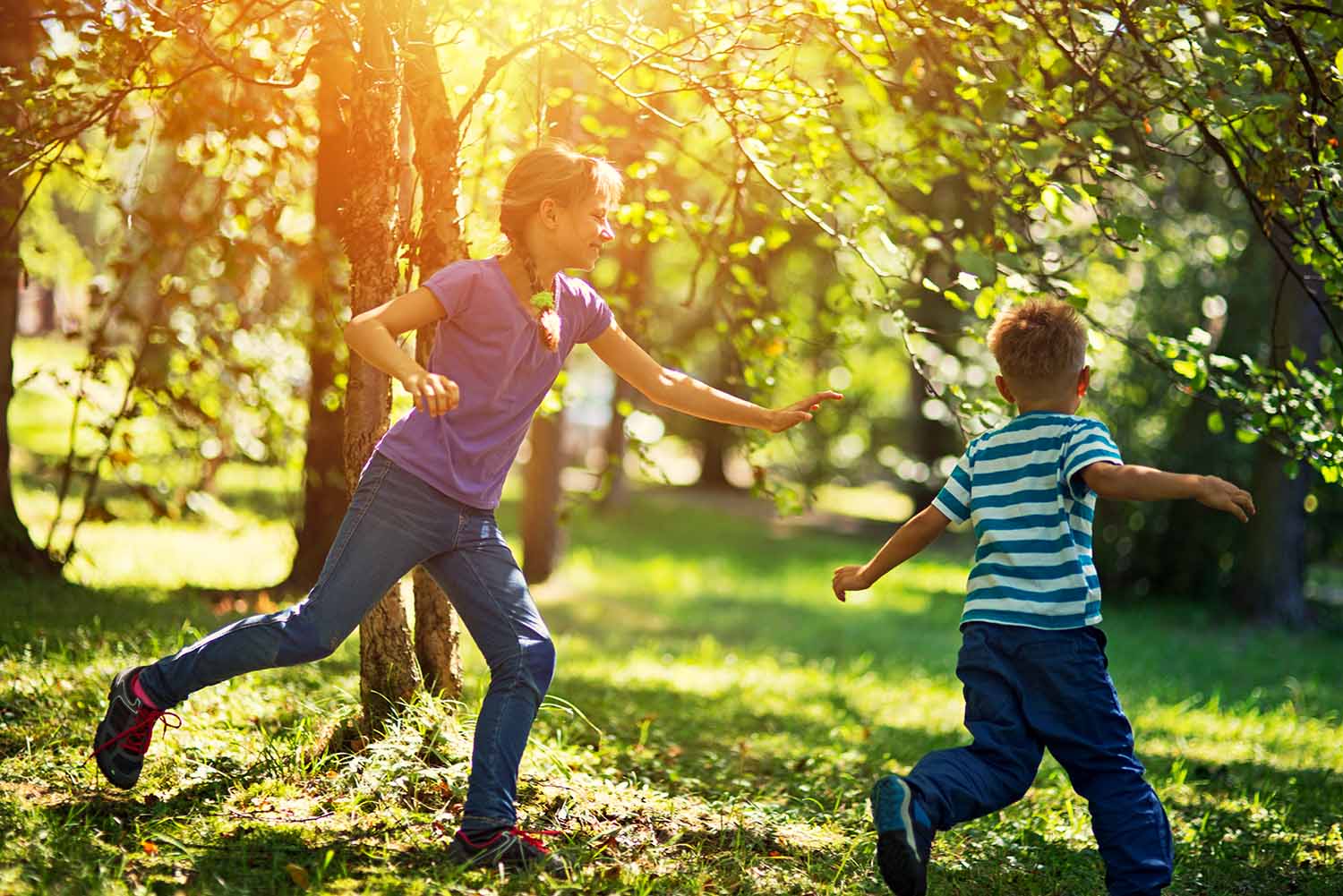 A boy and girl play tag among trees in a park.