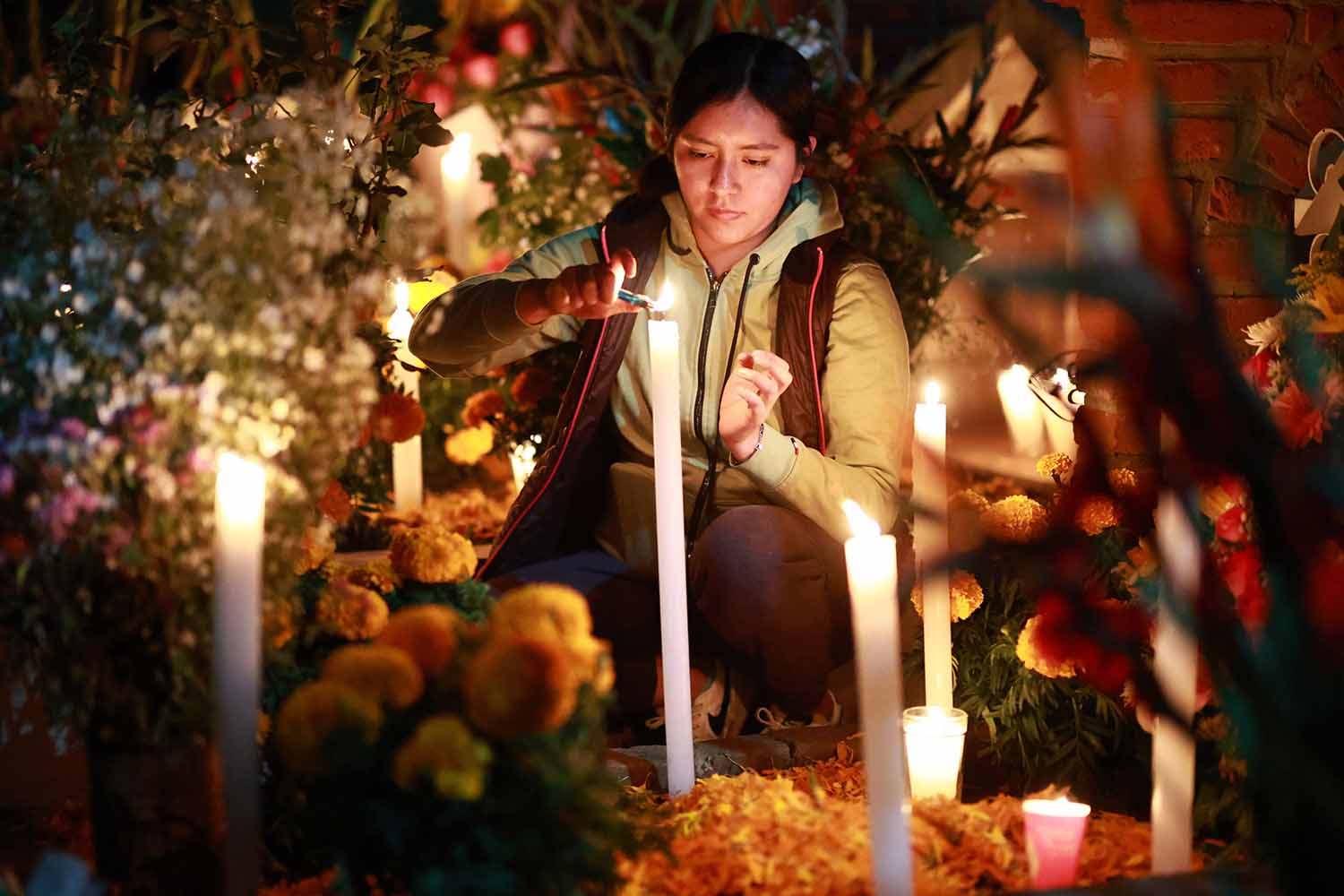 A woman is surrounded by flowers as she kneels and lights tall white candles outdoors at night.