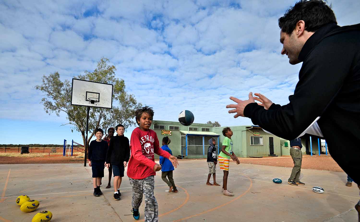 A man throws a ball to an Aboriginal child on a basketball court as other children watch.