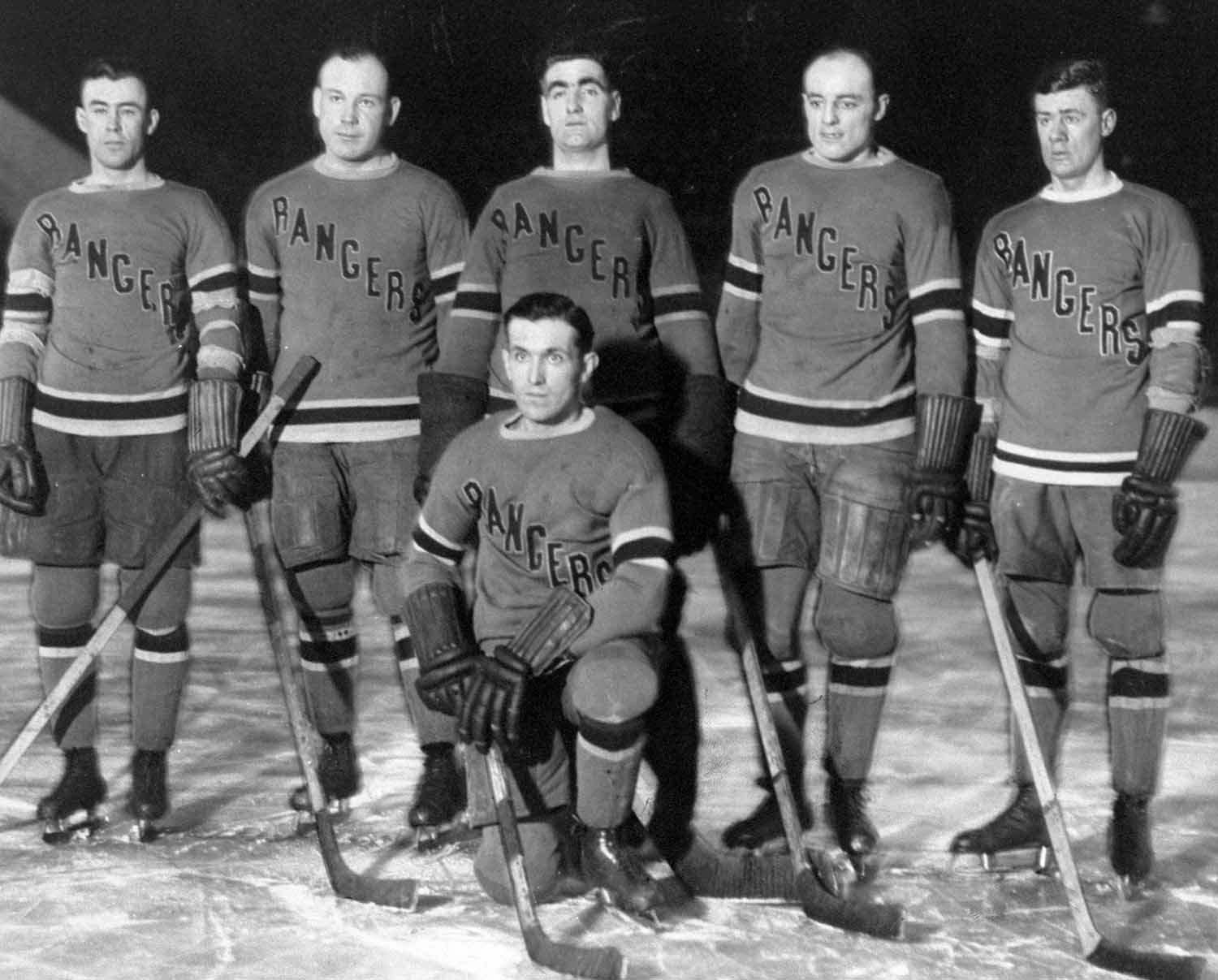 Five men in 1920s Rangers uniforms stand on their ice with their hockey sticks while a sixth man kneels in front.