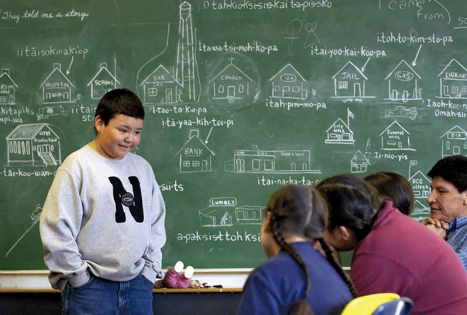 Children and an adult are in a classroom where a blackboard shows drawings of objects and the words for them in the Blackfeet Language.