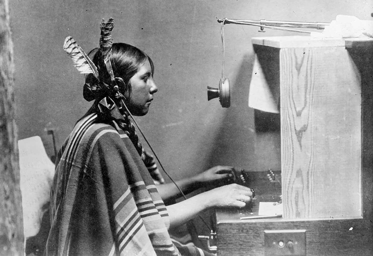 A woman in traditional Blackfeet clothing sits at a small telephone switchboard.