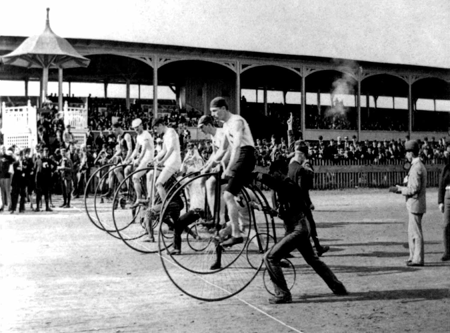A black and white image shows riders racing pennyfarthings in an arena with spectators watching.