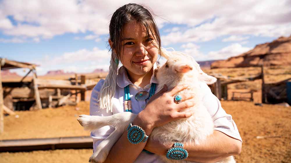 A young woman poses while holding a lamb with southwestern buildings and land features in the background.