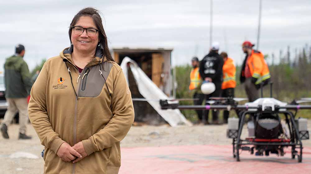 A woman poses outdoors in front of a large drone and some men in orange safety equipment.