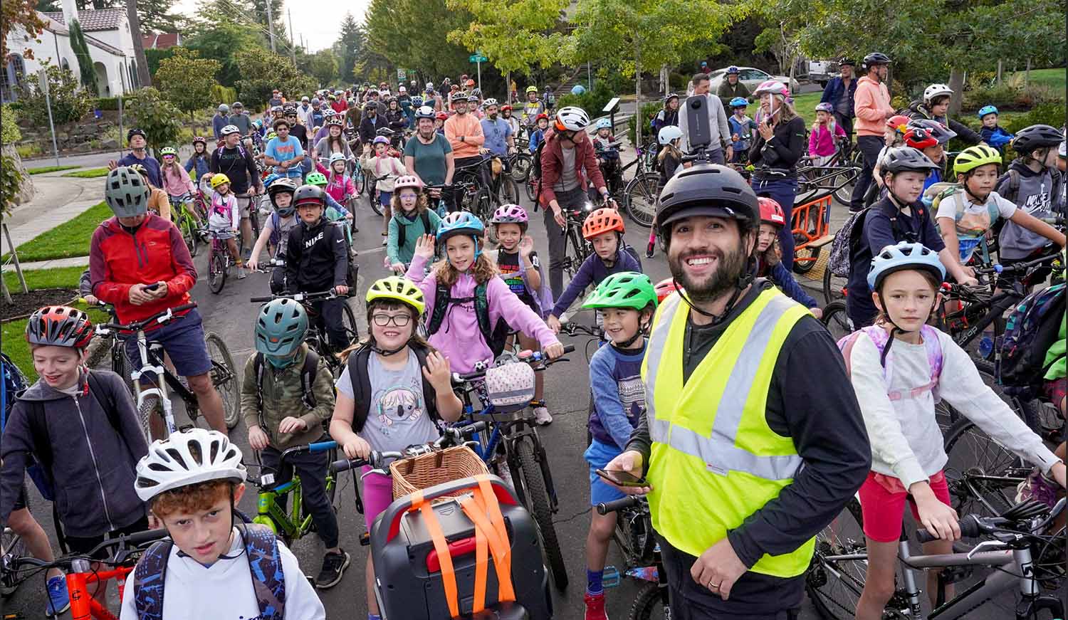 Sam Balto wears a neon safety vest and bike helmet as he poses with adults, children, and their bikes on a suburban street.