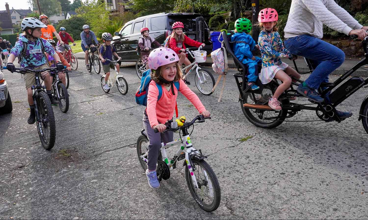 A group of adults and children of different ages ride their bikes down a suburban street.