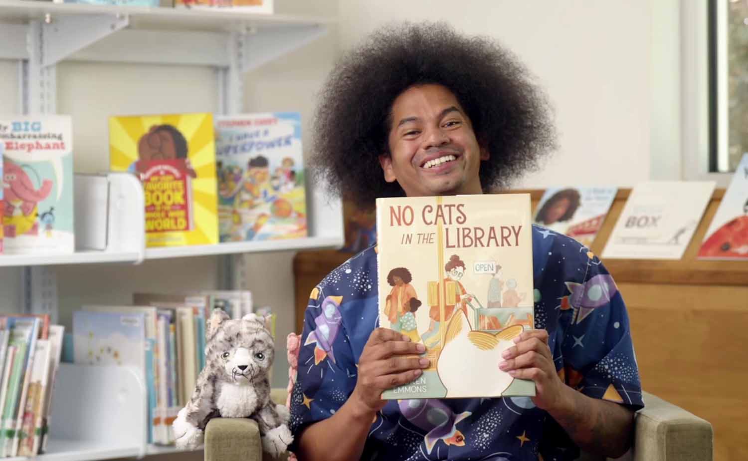 Mychal Threets poses in a children’s library while holding the book No Cats in the Library.