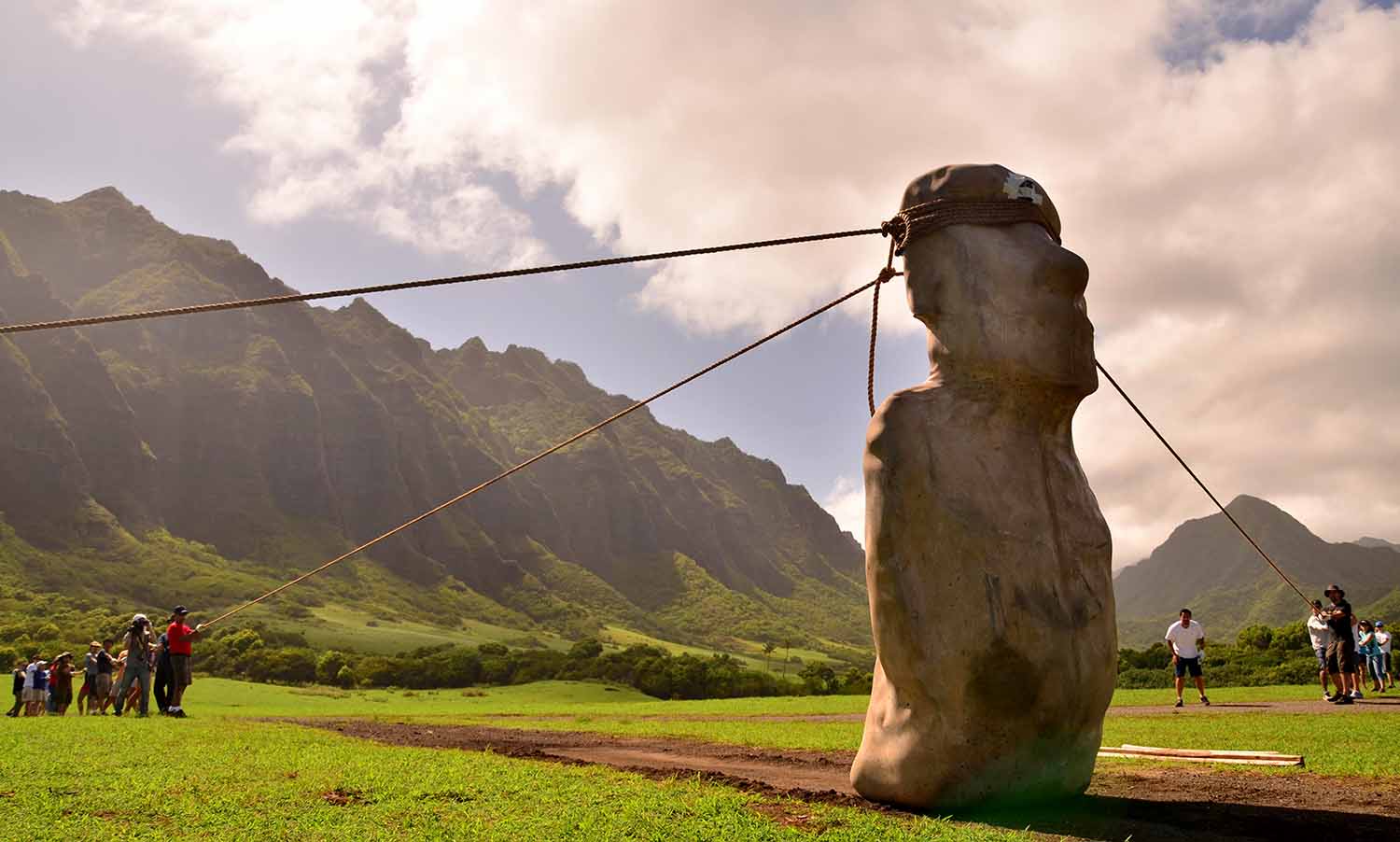 A replica of a large moai statue stands on a dirt path as people pull on ropes that are tied around its head.