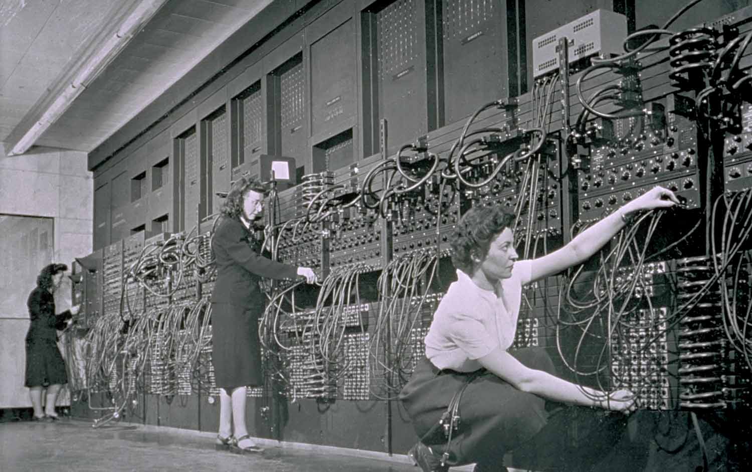 Three women manipulate the wires of a computer that runs the length of a large room.
