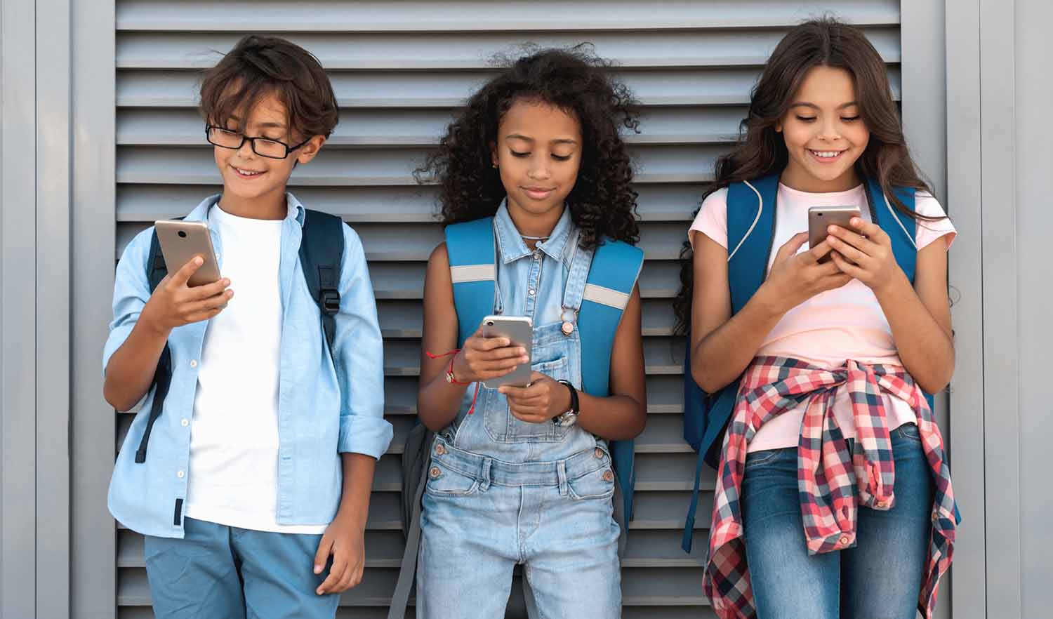 Three middle-school age kids stand against a wall looking at their phones.