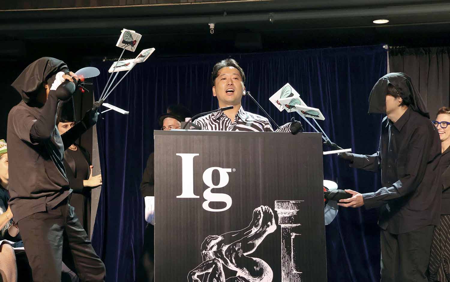 A man stands at a podium wearing a zebra patterned shirt while people in black hoods hold pictures of flies.