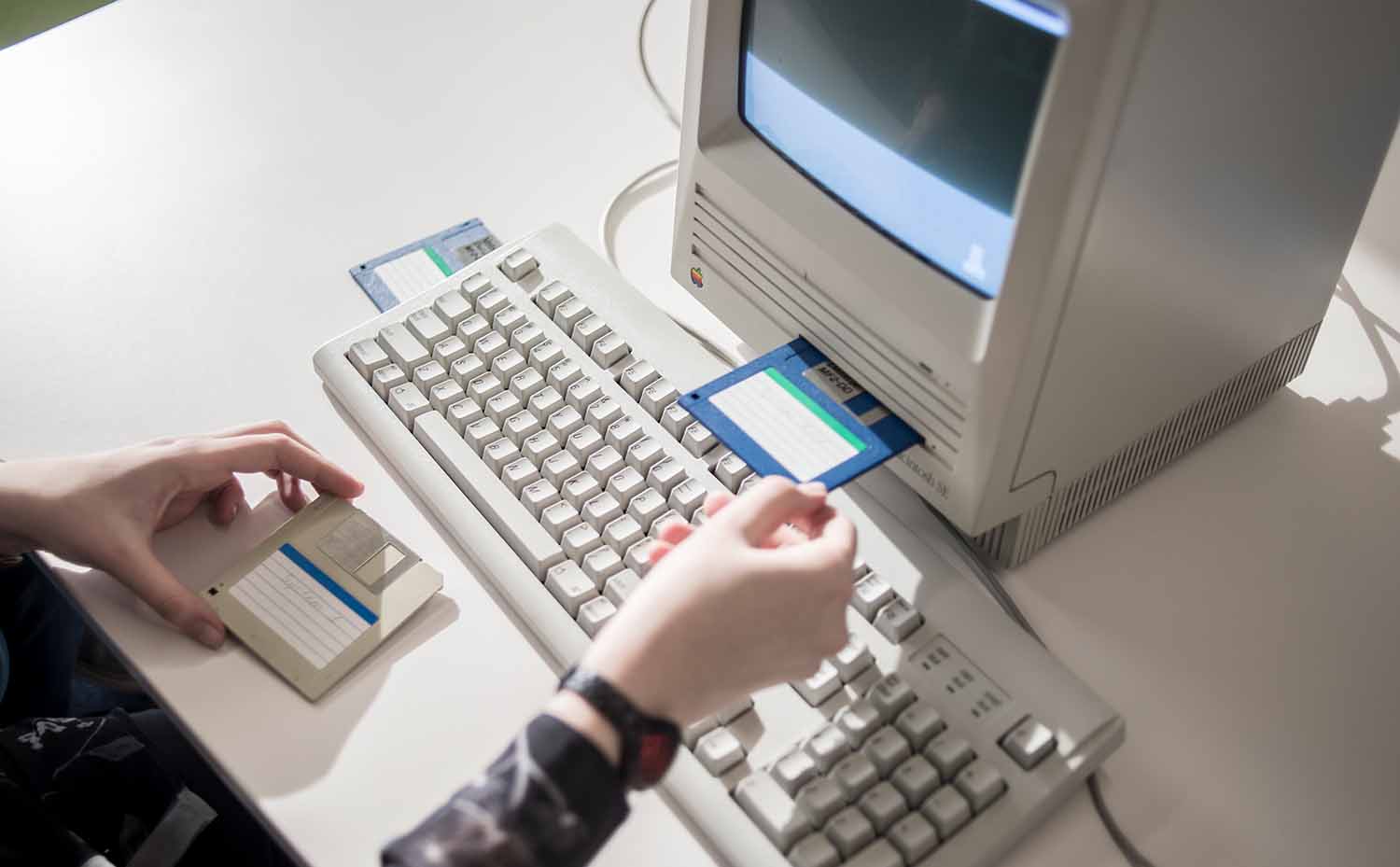 A person puts a three and a half inch floppy disk into the drive of a Mac computer while holding another disk.
