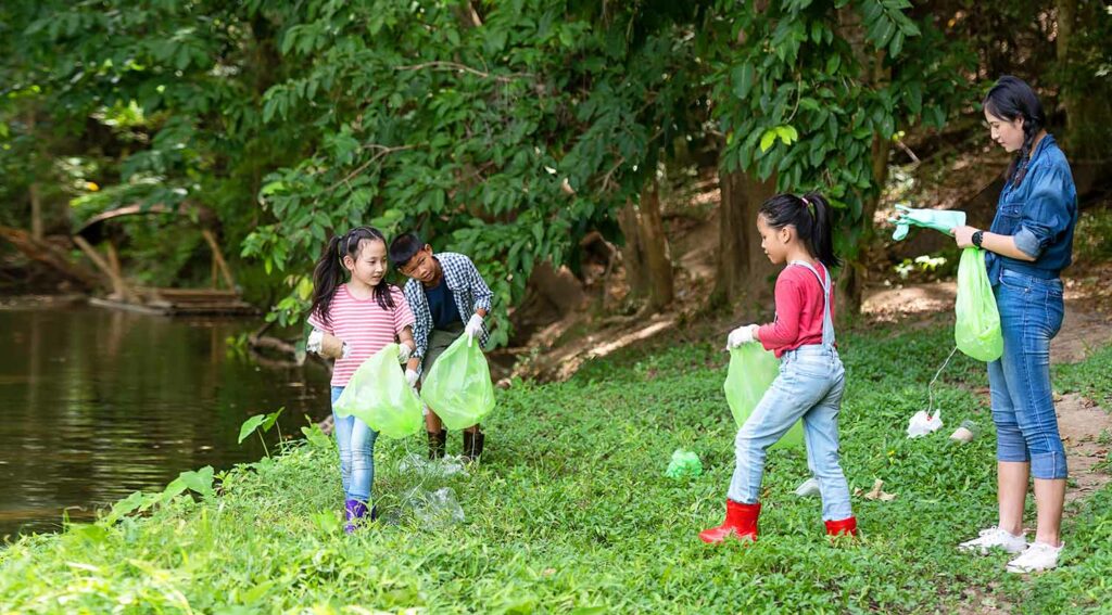 Three children and one adult stand in front of a body of water holding plastic bags and picking up plastic bottles.