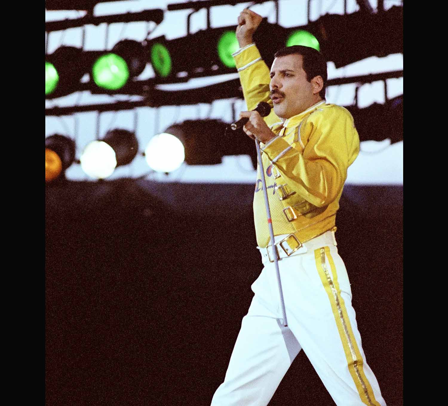 Freddie Mercury stands on stage holding a microphone in one hand and holding his other arm in the air.