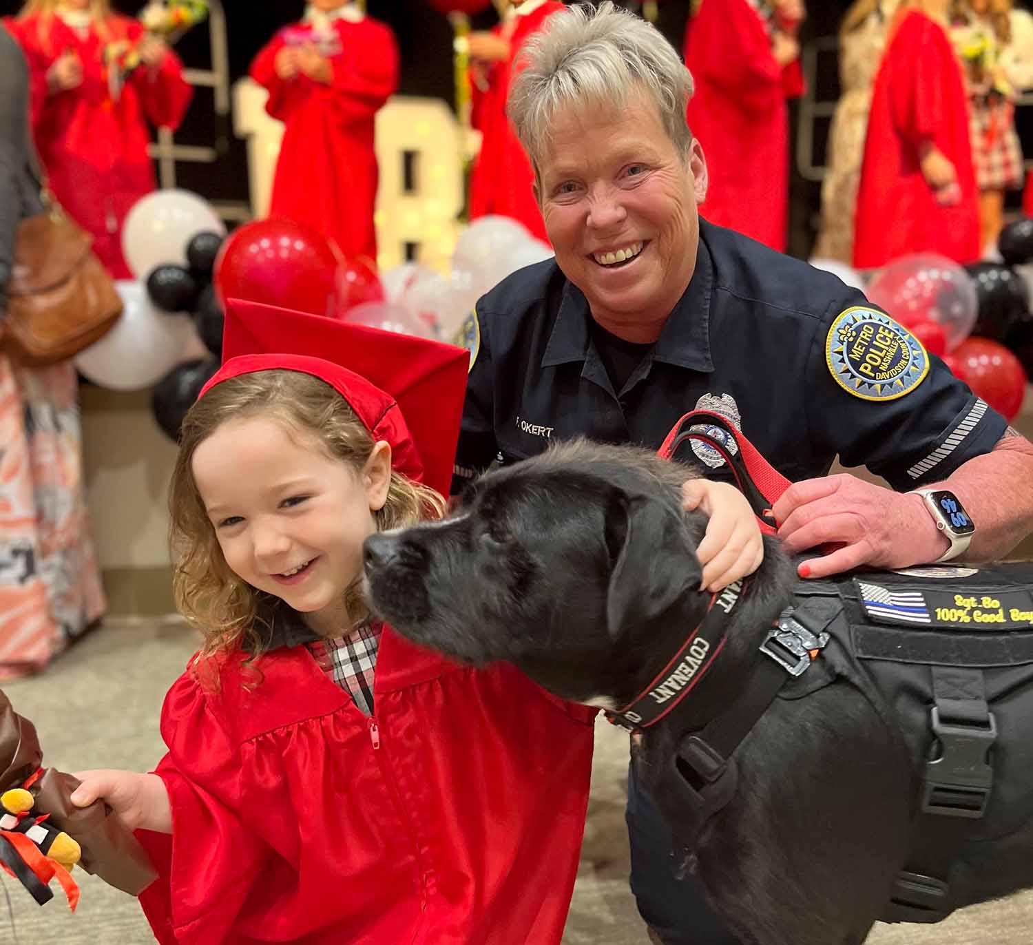 A police officer smiles and holds Bo’s harness as Bo sniffs the face of a young child at her graduation ceremony.
