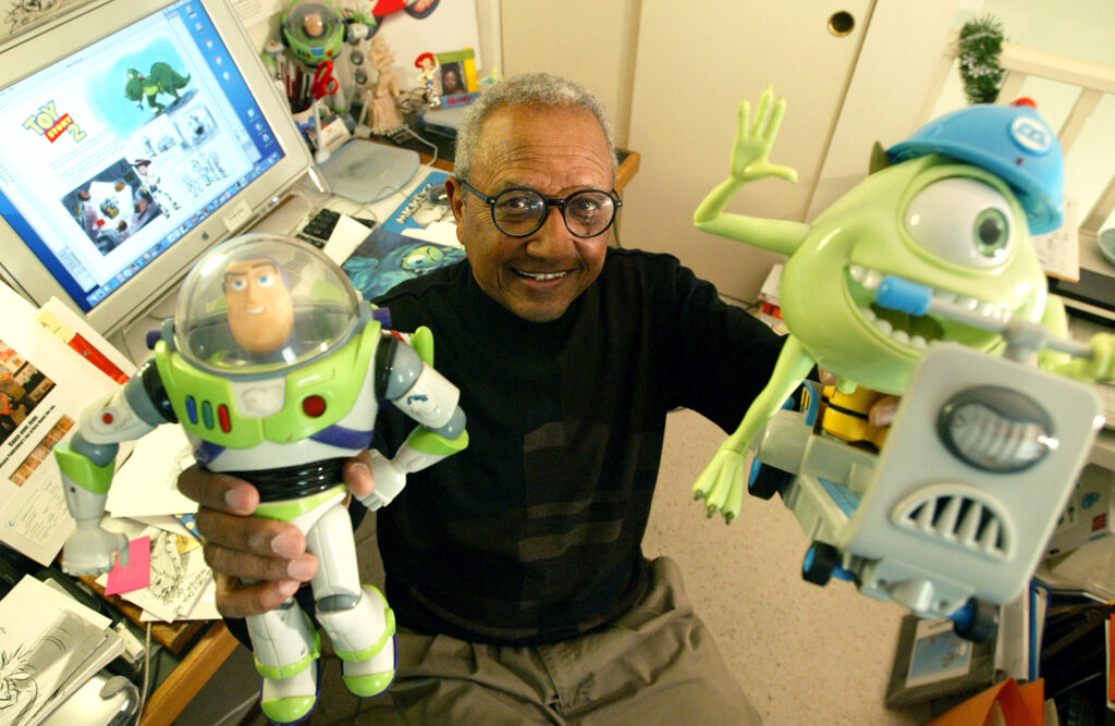 Animator Floyd Norman sits at a computer with the Toy Story logo on the screen and holds up Buzz Lightyear and Mike Wazowsky toys from the movies Toy Story and Monsters Inc.