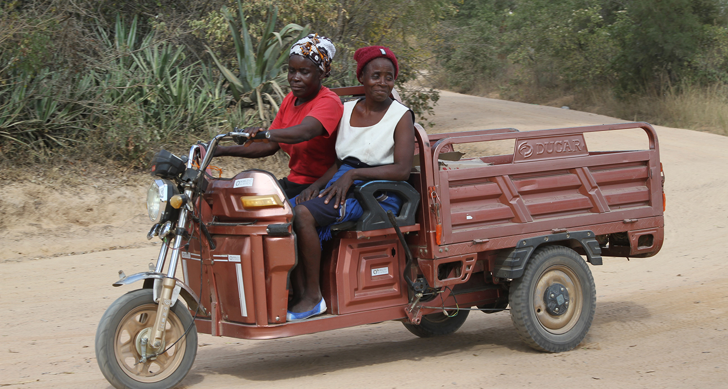 A woman and her passenger drive a three wheeled vehicle with an open cargo bed in the back.