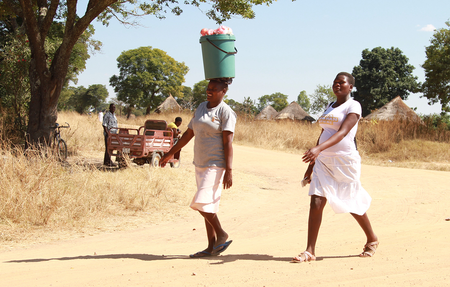 Two women walk side by side on a road, with one carrying a bucket of vegetables on her head and a three-wheeled vehicle with two men in the background.