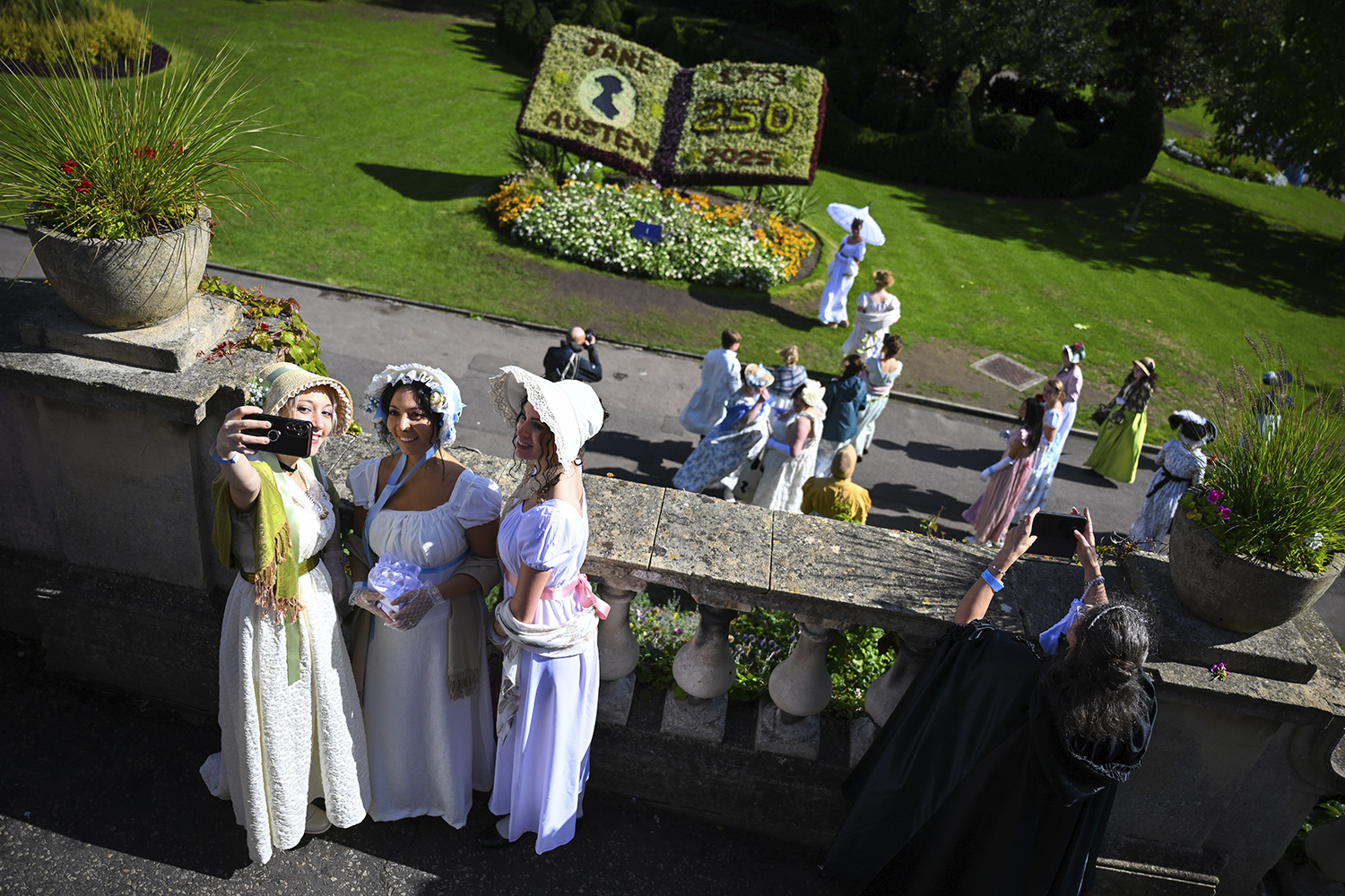Three women in Regency Era clothing take a selfie on a balcony. Behind them, a bush has been carved to resemble a book that says Jane Austen 250.