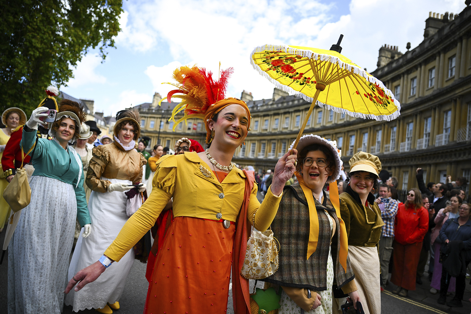 Women in Regency Era clothing smile as they walk down a street with a crowd and a historic building in the background.
