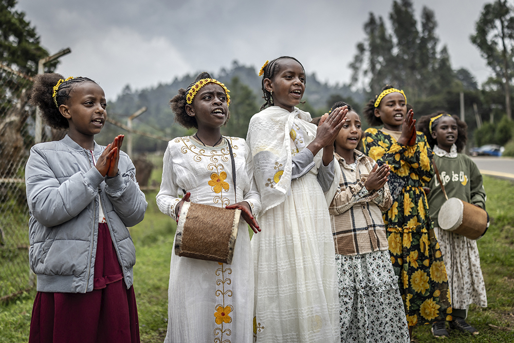 Six girls and young women wear floral-themed clothing as they stand outdoors singing.