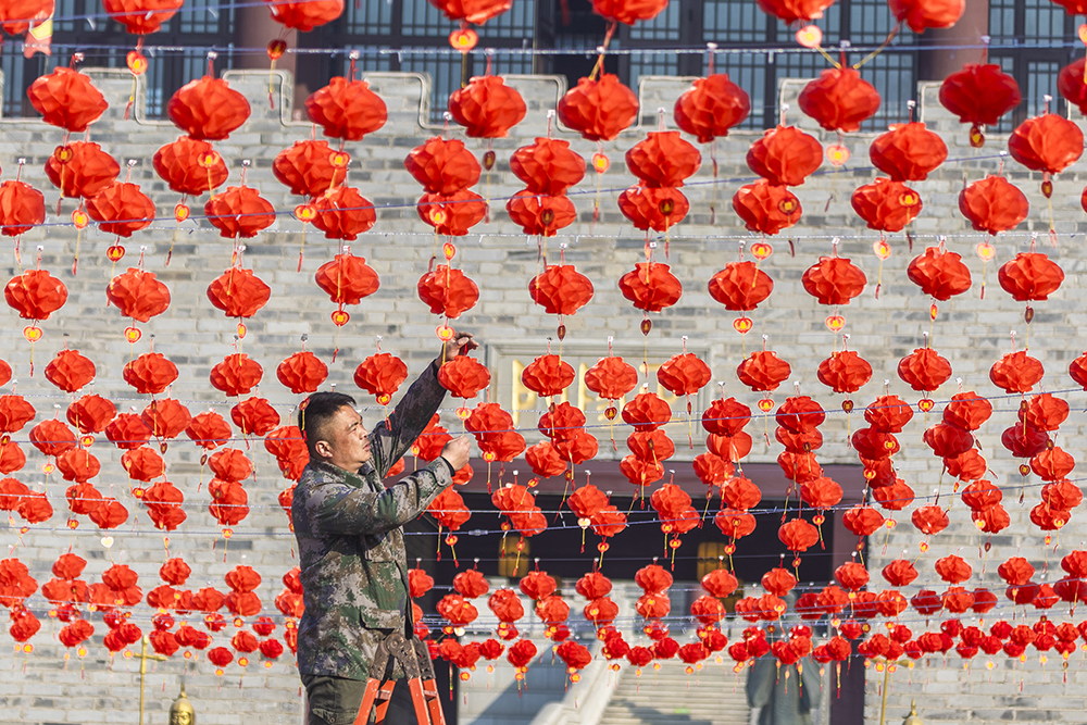 A man stands on a ladder and hangs multiple red lanterns in a plaza.