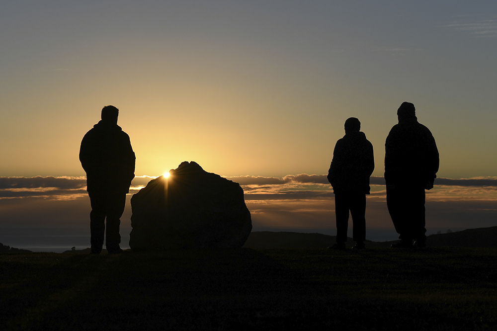 Three people and a large stone are silhouettes against a sunrise.