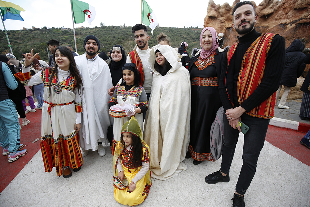 A family of nine smile as they pose for a photo outdoors in front of Algerian flags while wearing traditional clothing.