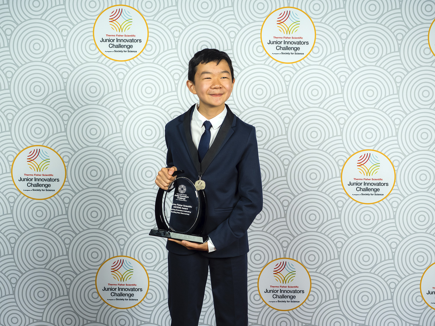 Miles Wu smiles and holds a trophy in front of a backdrop reading Junior Innovators Challenge