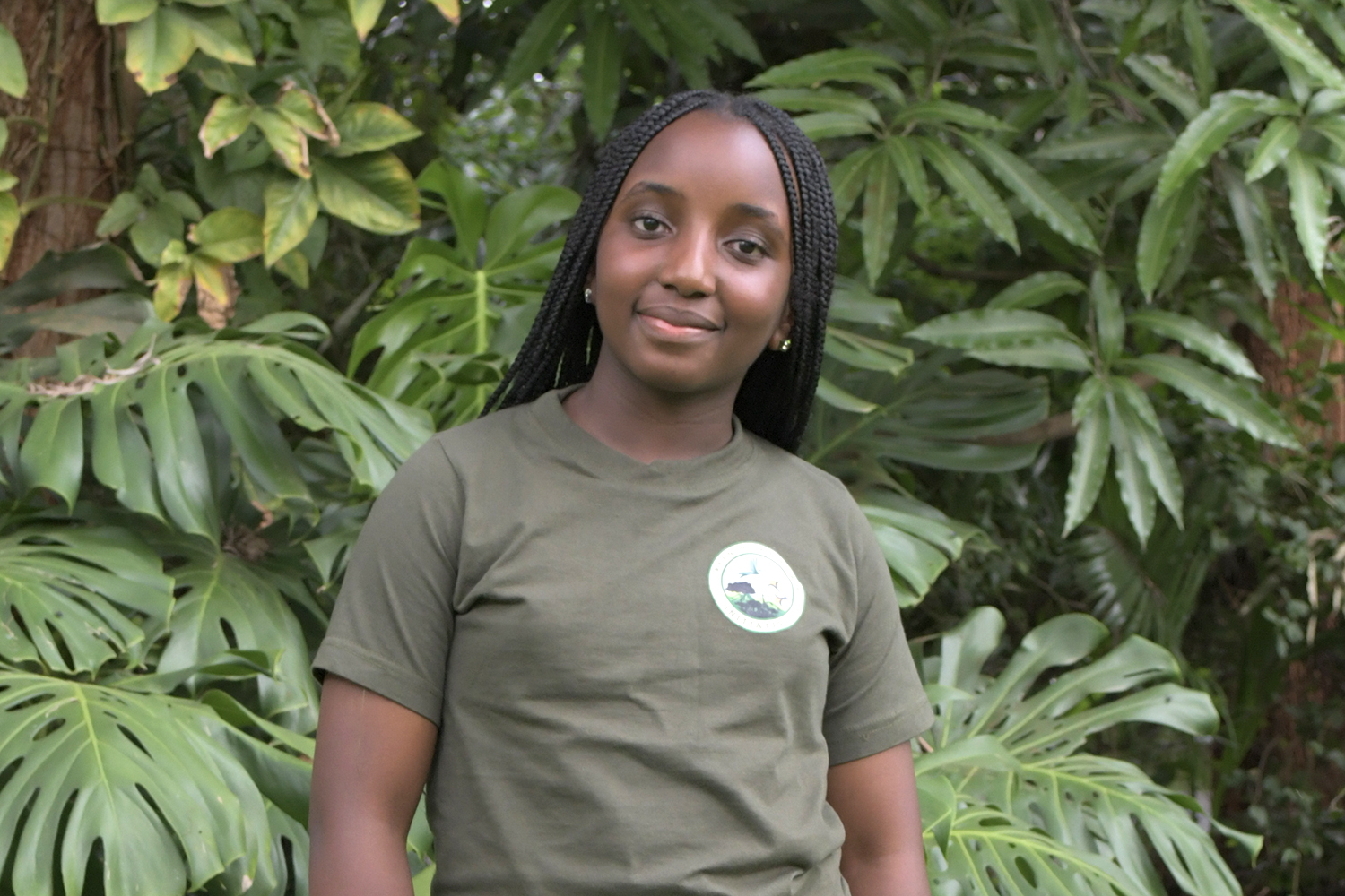 A headshot of Alice Wanjiru shows her wearing a tee shirt and posing in front of greenery.