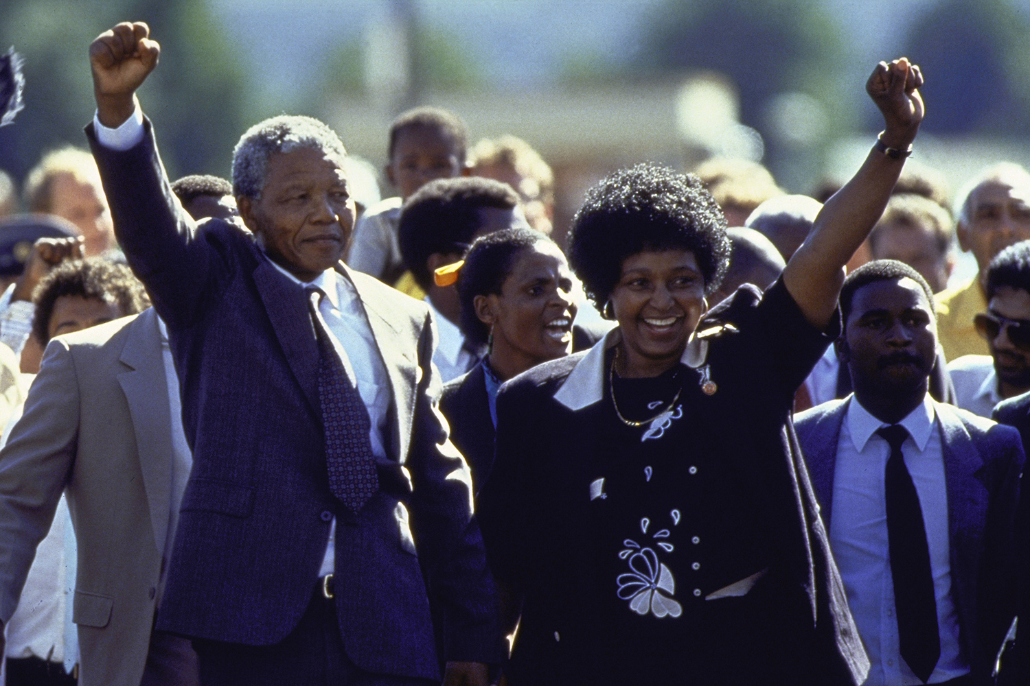 Nelson and Winnie Mandela each hold one arm up and smile with a crowd in the background.