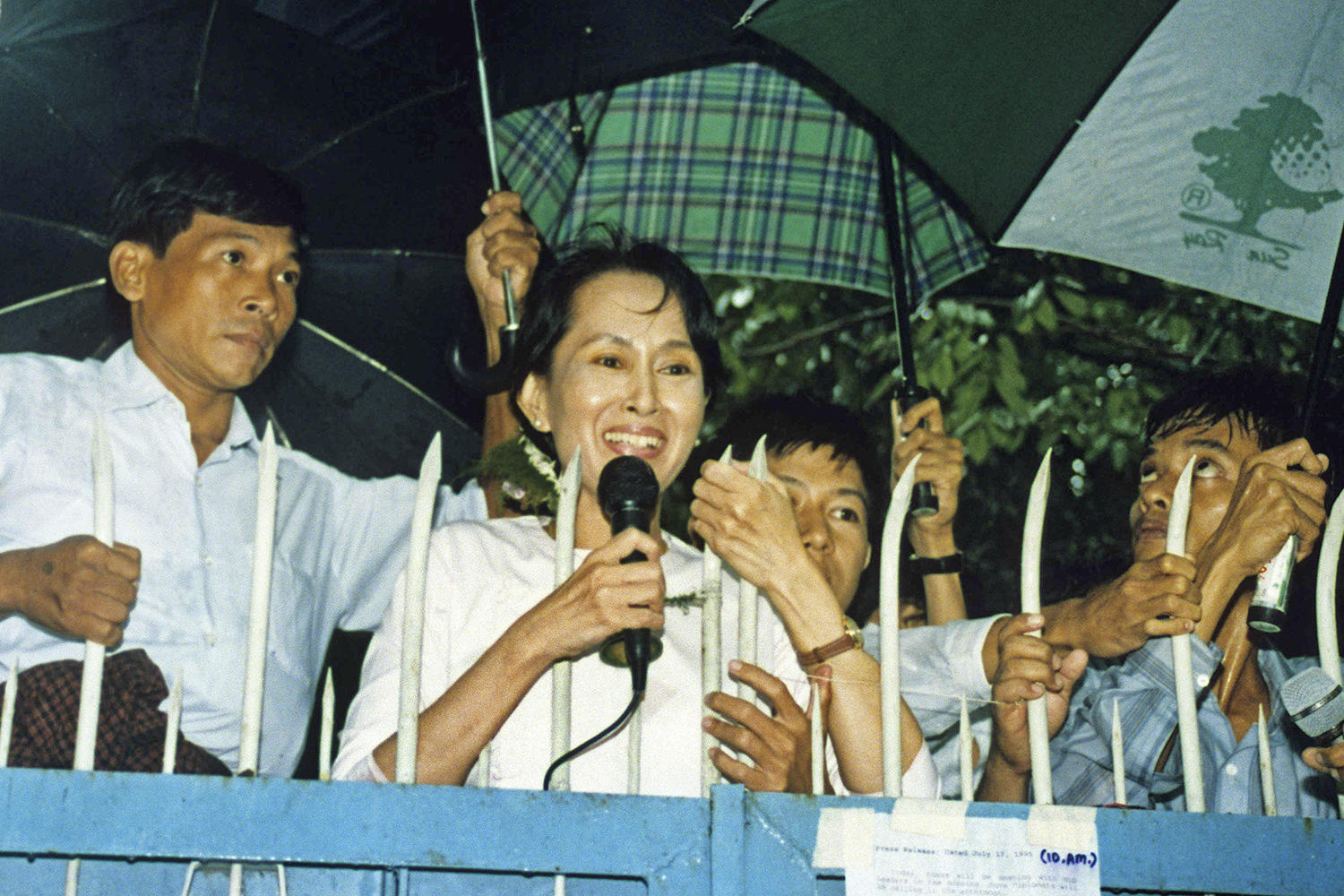 Aung San Suu Kyi smiles as she holds a microphone to speak while several people hold umbrellas behind her.