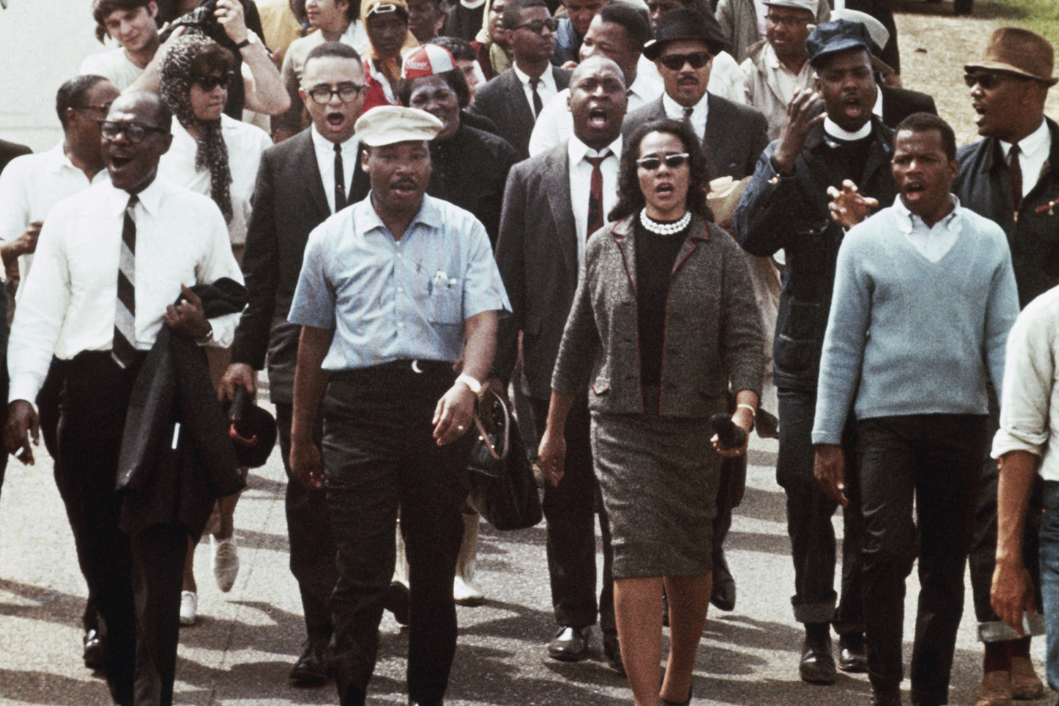 Martin Luther King and Coretta Scott King lead a group of activists, some carrying US flags, as they march down a street.