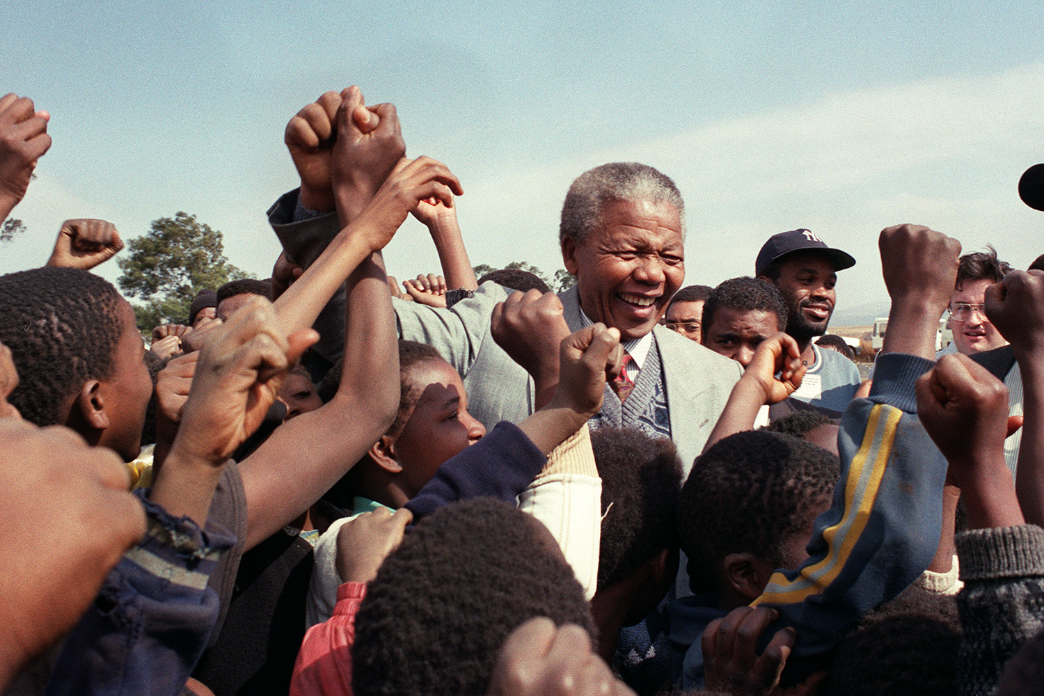 Nelson Mandela stands in a group of children and adults and smiles while grabbing the hand of one person in the crowd.