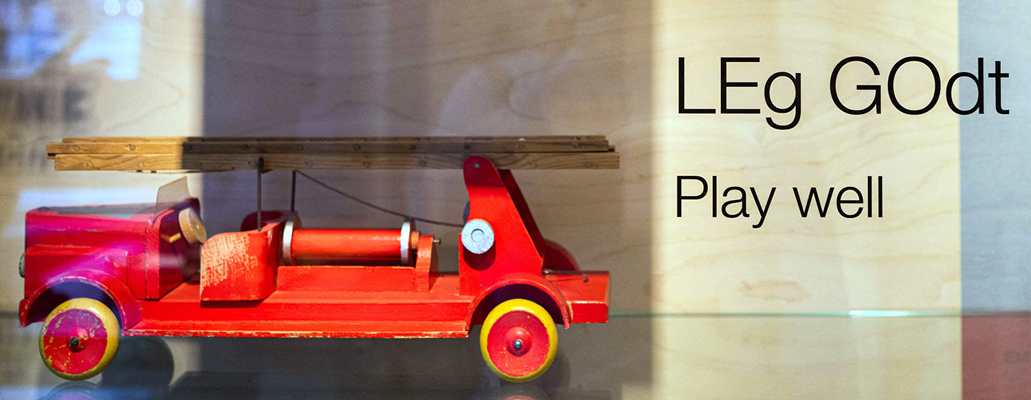A wooden toy fire truck is in a display case next to the words LEG GODT and play well.