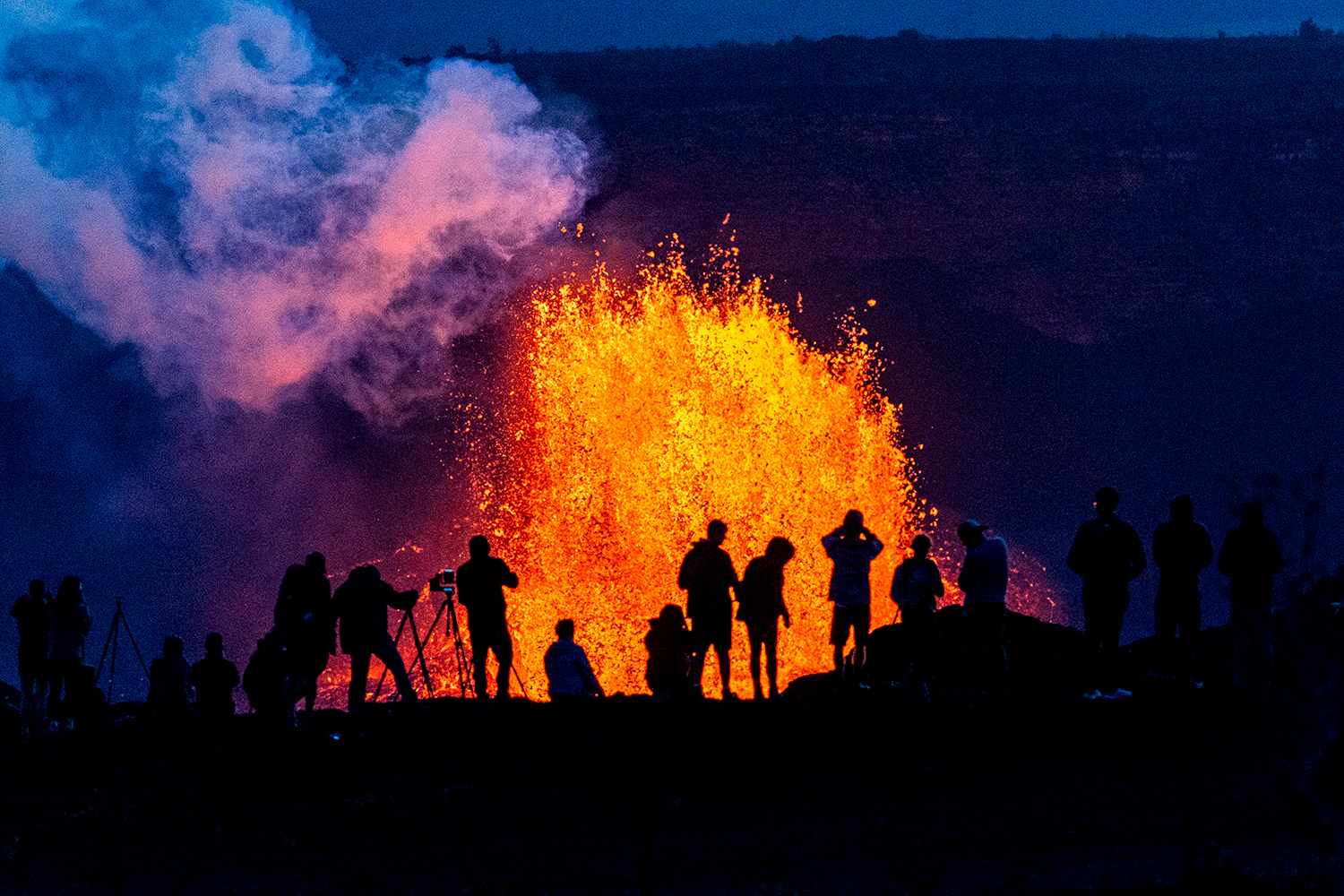 The silhouettes of several people and a camera on a tripod are seen against lava that is spewing into the air at night.